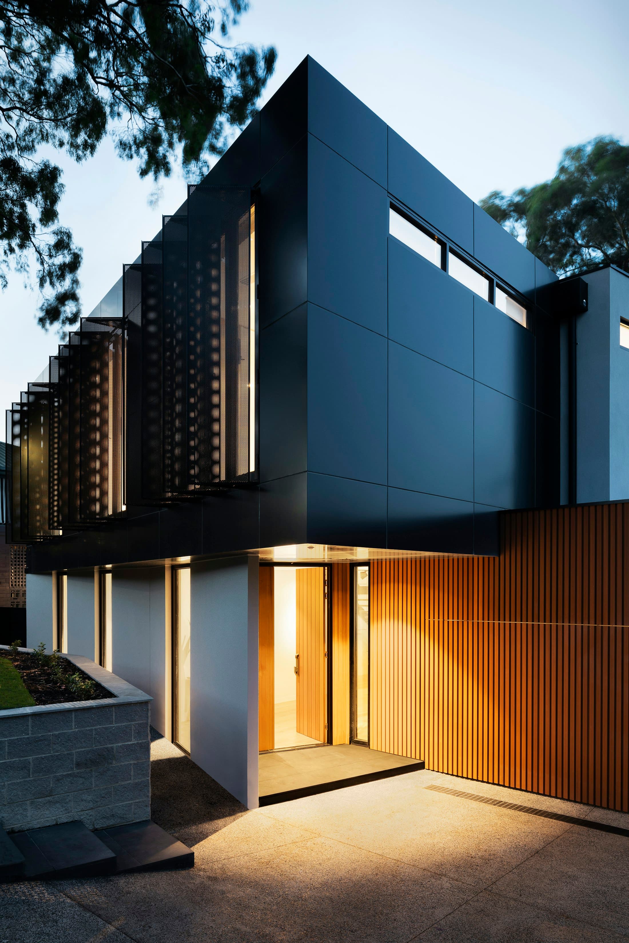 The entry court at nightfall. Vertical timber slats filter light from the hall, marking the threshold without a single sign.